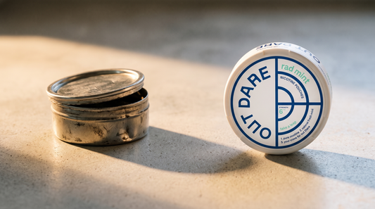 Worn unbranded snus can beside a clean round OutDare nicotine pouch can on a concrete worktop