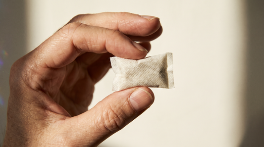 An adult hand pinching a single small flat white nicotine pouch sachet under raking sidelight revealing soft textile texture