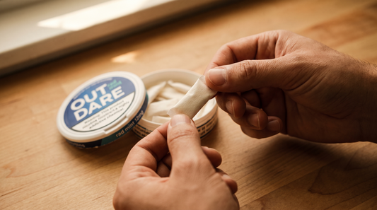 Close-up of adult hands examining a small white nicotine pouch with an open can softly visible behind on warm wood