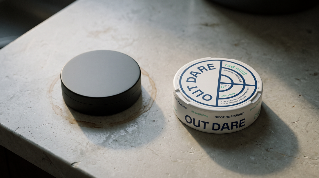 A worn dark snus can beside a clean OutDare nic pouch can on pale stone