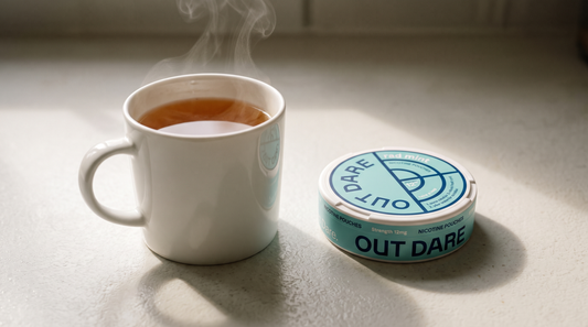 Mint-green nicotine pouch can beside a steaming mug of amber tea on a pale kitchen worktop