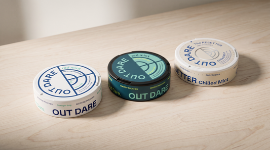 Three OutDare nicotine, energy and sleep pouch cans lined up on a pale birch-wood surface