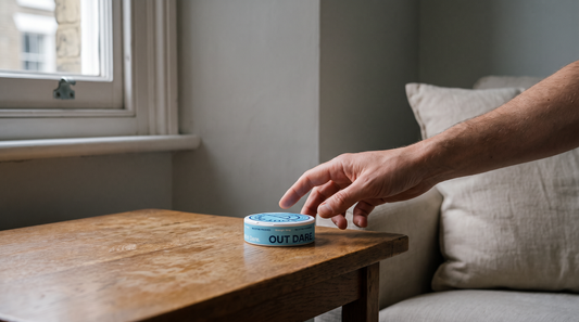 Adult hand reaching toward a nicotine pouch can on a worn oak table in a British interior