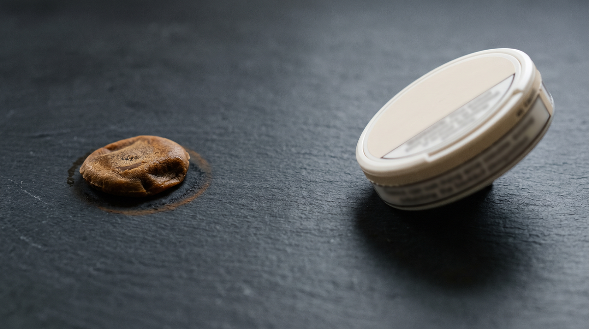 Brown used snus portion and closed nicotine pouch can on a dark slate surface