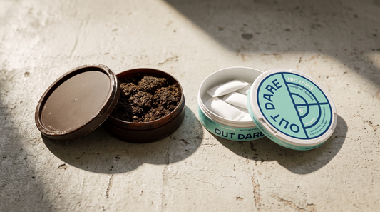 Dark matte snus can open beside an OutDare nikotino maišeliai can on a concrete surface