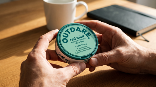 Adult hands examining a round teal OutDare Rad Mint nicotine pouch can above a wooden desk