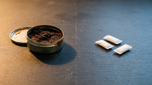 Open tobacco can with brown residue beside clean white nicotine pouches on a slate surface